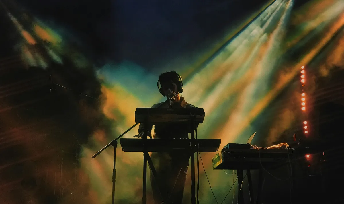 Musiker am Keyboard als Silhouette vor goldenen und türkisen Bühnen-Lichtkegeln im Nebel.