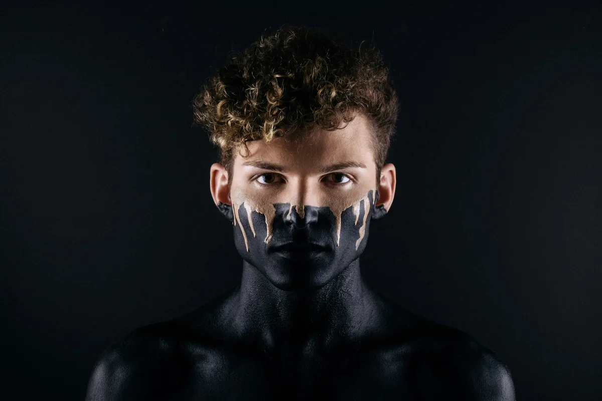 Ausdrucksstarkes Editorial-Porträt eines jungen Mannes mit schwarzer Gesichtsbemalung vor dunklem Hintergrund, direkter Blick.